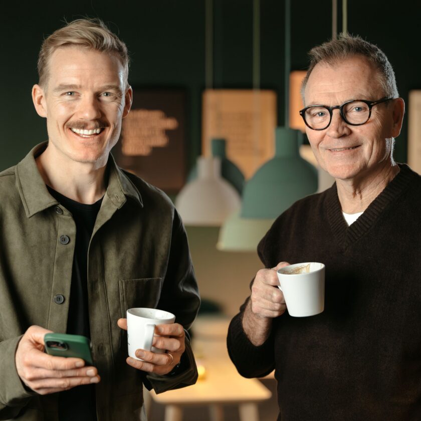 Nordlo. Två män dricker kaffe och tittar in i kameran.